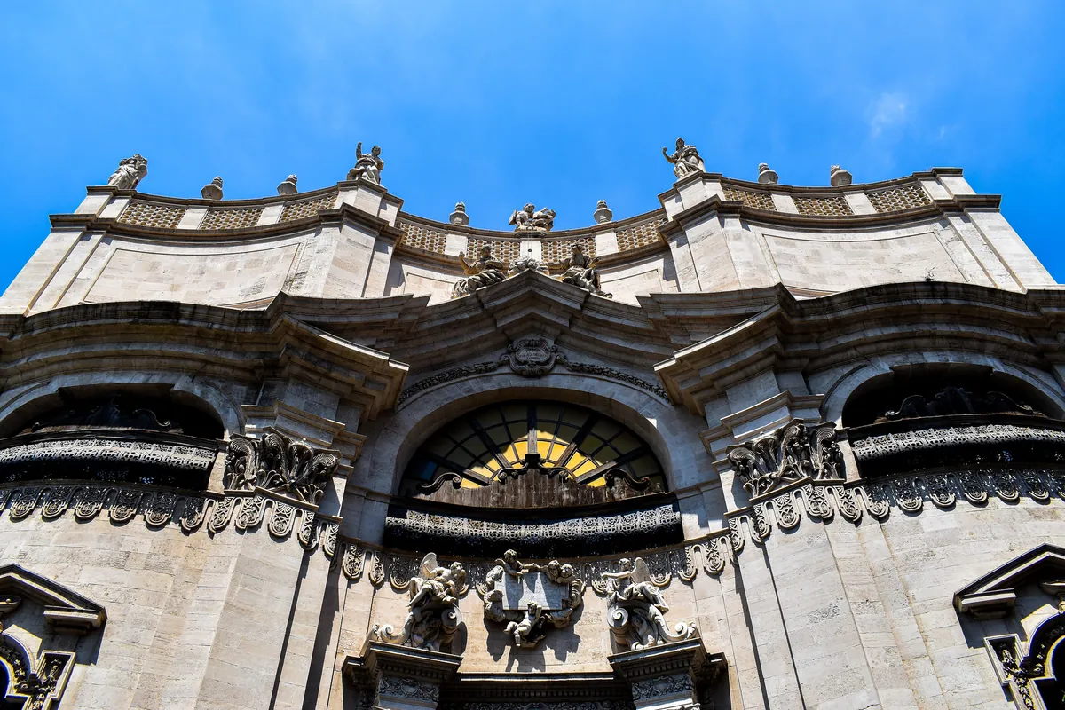 Zakřivená kamenná fasáda Badia di Sant'Agata pohledem zdola, s vyřezávanými římsami, sochami a velkým oknem nad vchodem na pozadí modré oblohy.