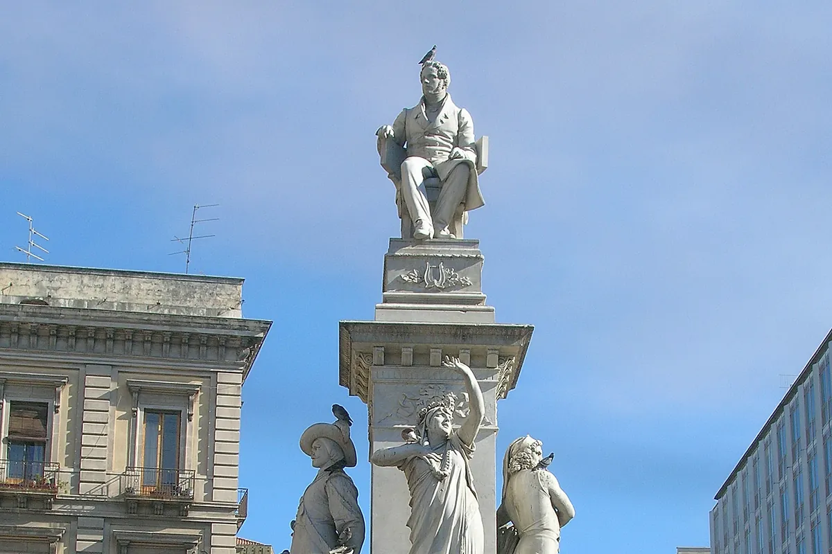 Belliniho pomník na Piazza Stesicoro v Katánii, bílá mramorová postava skladatele sedícího na vysokém podstavci se sochami kolem základny pod modrou oblohou