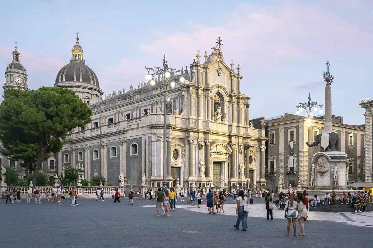 Piazza del Duomo v Catanii za soumraku, s fasádou katedrály, fontánou slona z lávového kamene a jejím obeliskem a lidmi přecházejícími náměstí.
